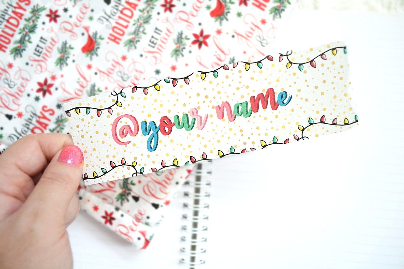 Image of a customizable bookmark being held in front of wrapping paper. The bookmark features holiday lights, and the words "@ your name." It is being held by a white hand with pink fingernail polish. 