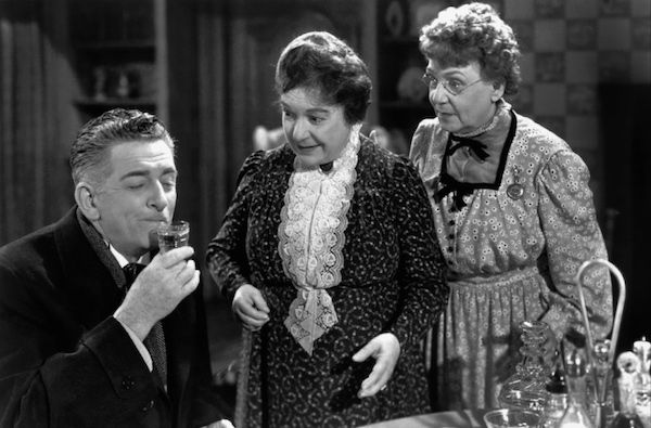 A black and white image of a seated man who begins to sip a cordial.  Aunts Abby and Martha look on excitedly to see the results. 