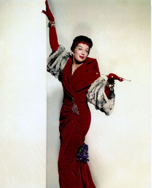 Rosalind Russell is wearing a cherry red velour coat with fur sleeves along with matching red gloves and headband. She carries a cigarette holder in her left hand and leans elegantly on a corner with her right. 