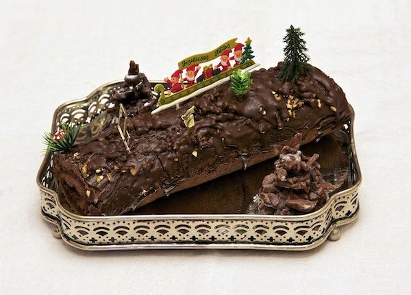 A bûche de Noël on a gold platter. The log is decorated with pine trees and plastic mushrooms, and has a sign on top that says "Joyeuses fêtes." 