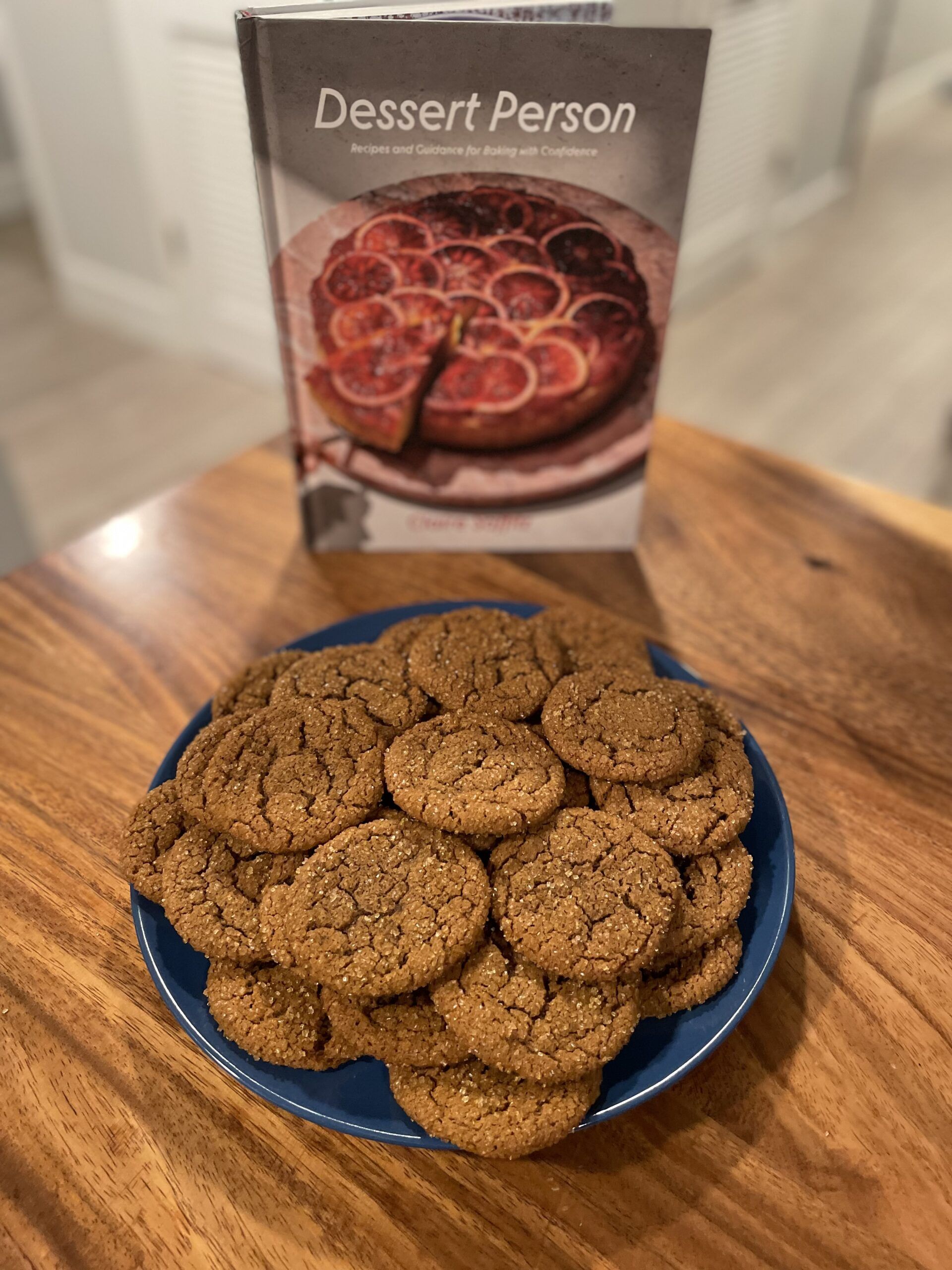 Plate of thin, sugar-coated ginger molasses cookies on a blue plate with a copy of Claire Saffitz's cookbook Dessert Person