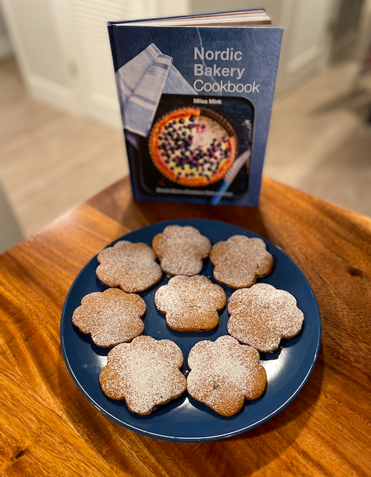 Gingerbread cookies shaped like puppy paws dusted with powdered sugar on a blue plate, alongside Miisa Mink's Nordic Bakery Cookbook