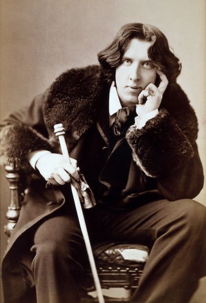 A sepia photograph of Oscar Wilde from 1882, at age 28. He is seated, leaning forward onto his knee, holding a cane. He wears a suit and a jacket with fur-trimmed collar and sleeves. 