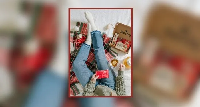 An overhead view of a person in jeans and a sweater holding a cup of tea while sitting on a white bedspread with a green and red tartan blanket and white ornaments scattered around. A box from SIPS BY, full of assorted bags of tea, sits open next to them.