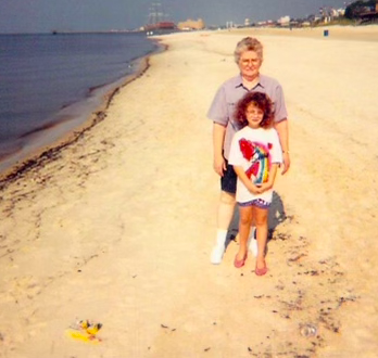 Photograph from 1990s on a beach of young girl with curly hair, standing in front of her grandmother with gray hair and glasses