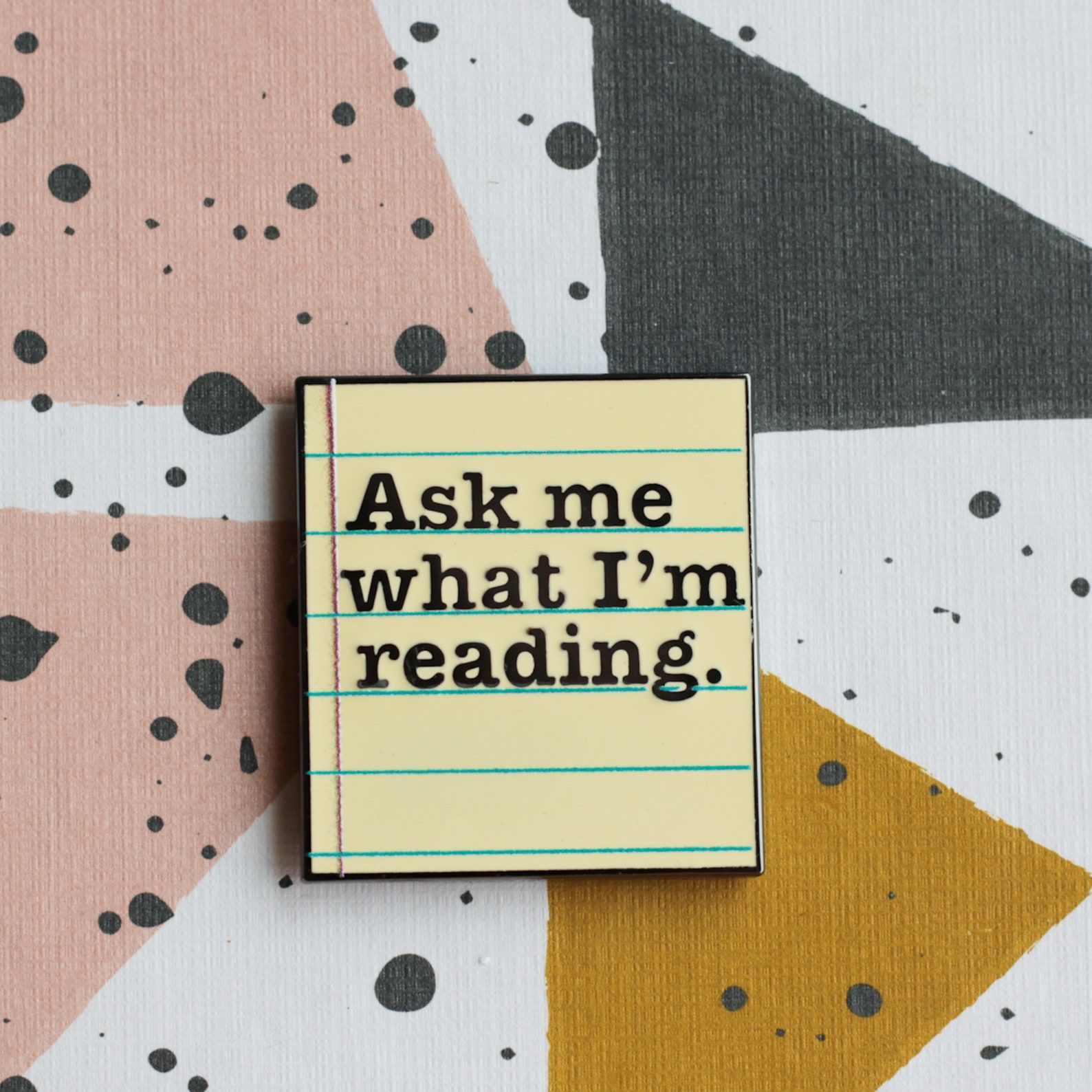 Image d'une épingle en émail jaune sur fond coloré.  La broche lit "Demandez-moi ce que je lis." 