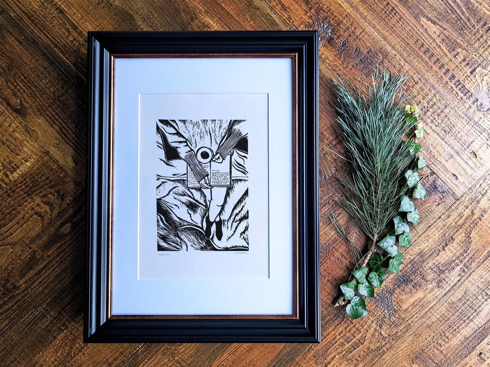Image of a linocut print that features a person reading with a cup of tea. It's in a black frame on a wooden background. 