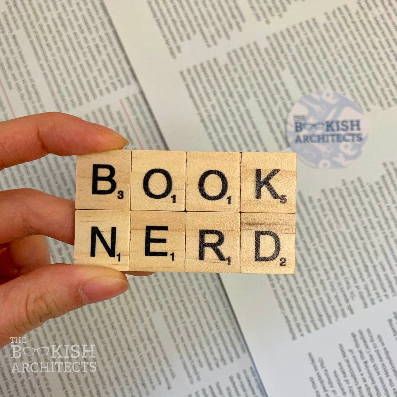 Image of a magnet made of Scrabble letters. The top row reads "book" and the bottom reads "nerd." It's being held by a white hand over open book pags. 