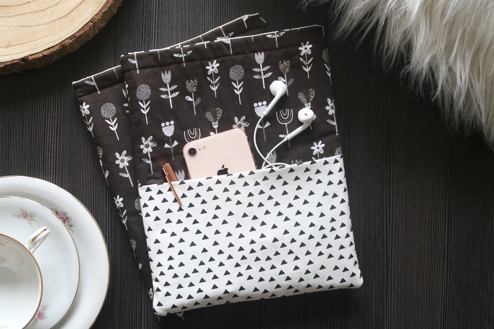 Image of a black and white fabric book sleeve on a black table, beside a tea cup. The sleeve has a white layer of fabric with black triangles and a black layer of fabric with whit flowers. 