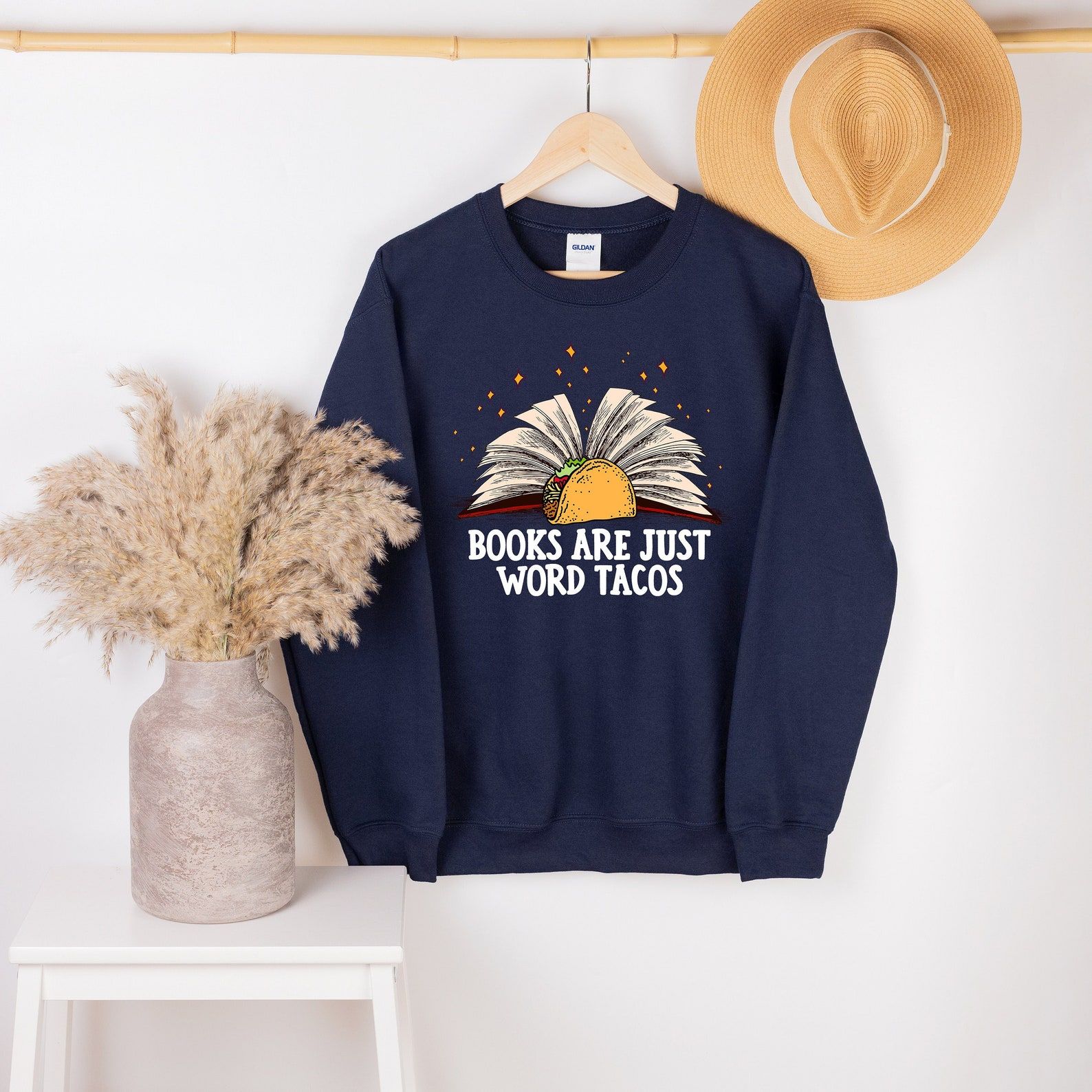 Navy sweatshirt hanging on a white wall. The sweatshirt has white text which reads "books are just word tacos," with a taco above it and a book behind the taco. The book is open. 