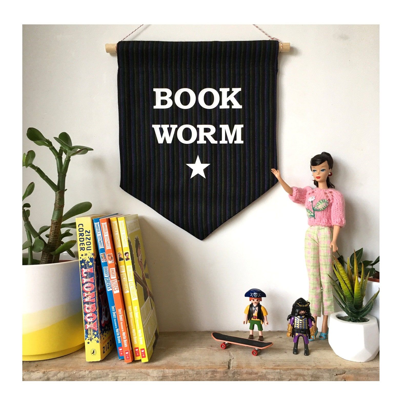 Black banner reading "Book worm" in white font with a white star. It is hanging above a bookshelf with books, plants, and children's toys. 