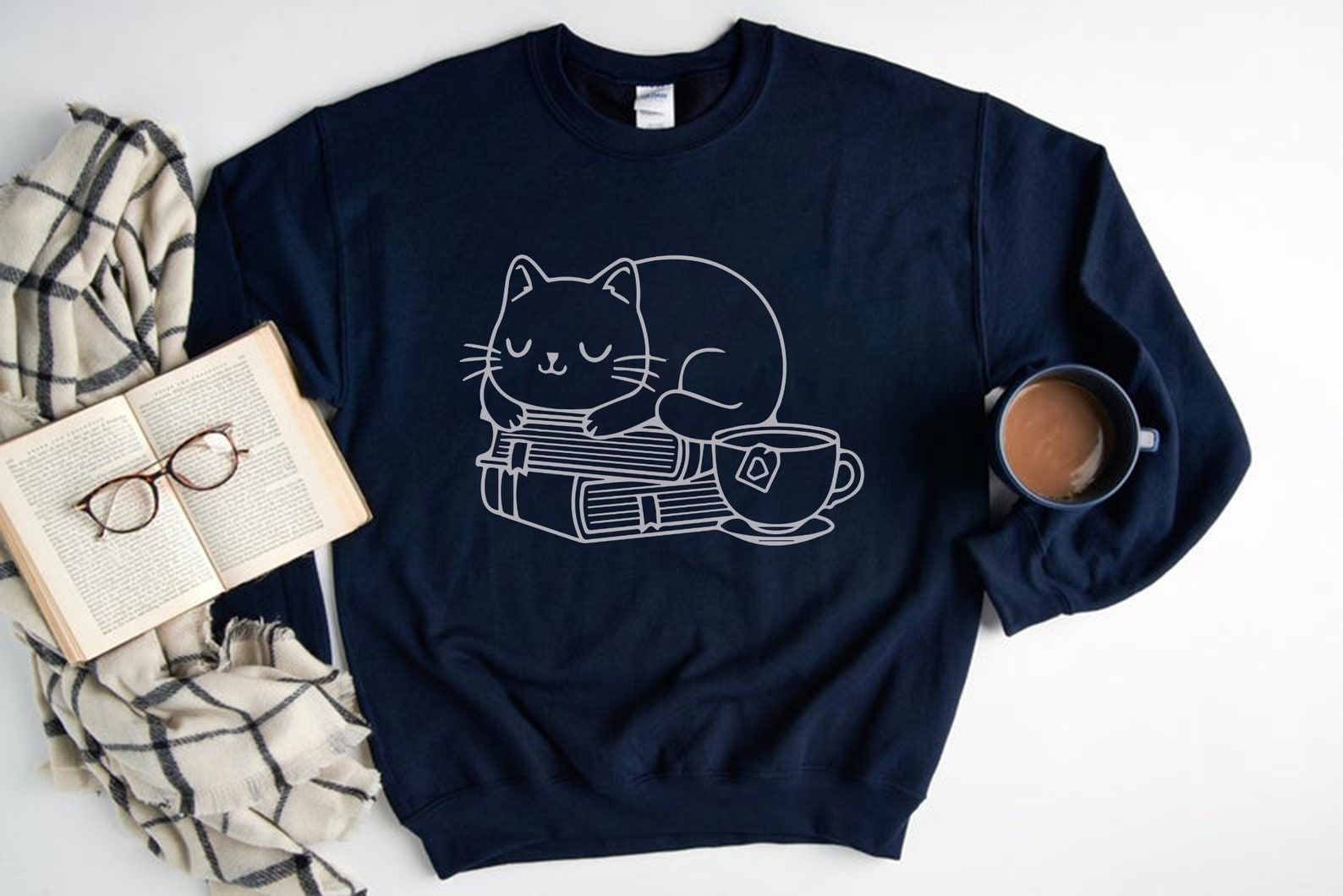 Blue sweatshirt with a white outline of a cat, cup of tea, and a stack of books. It is beneath an open book that has glasses on top, as well as with a mug of coffee under the sleeve's seam. 