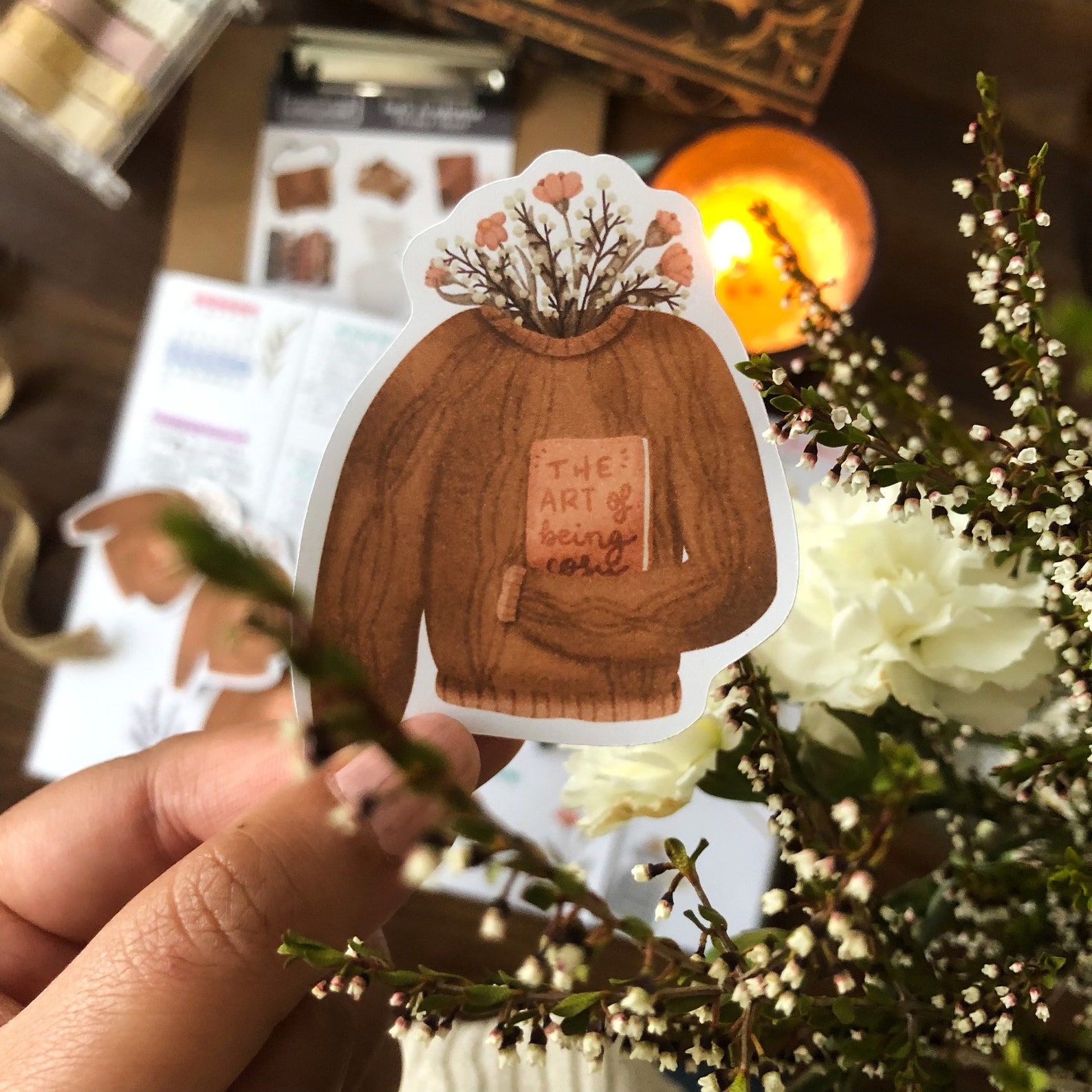 Image of a brown hand holding a sticker. The sticker is a brown knit sweater with a book tucked under the sleeve. The collar has flowers bursting from it. 