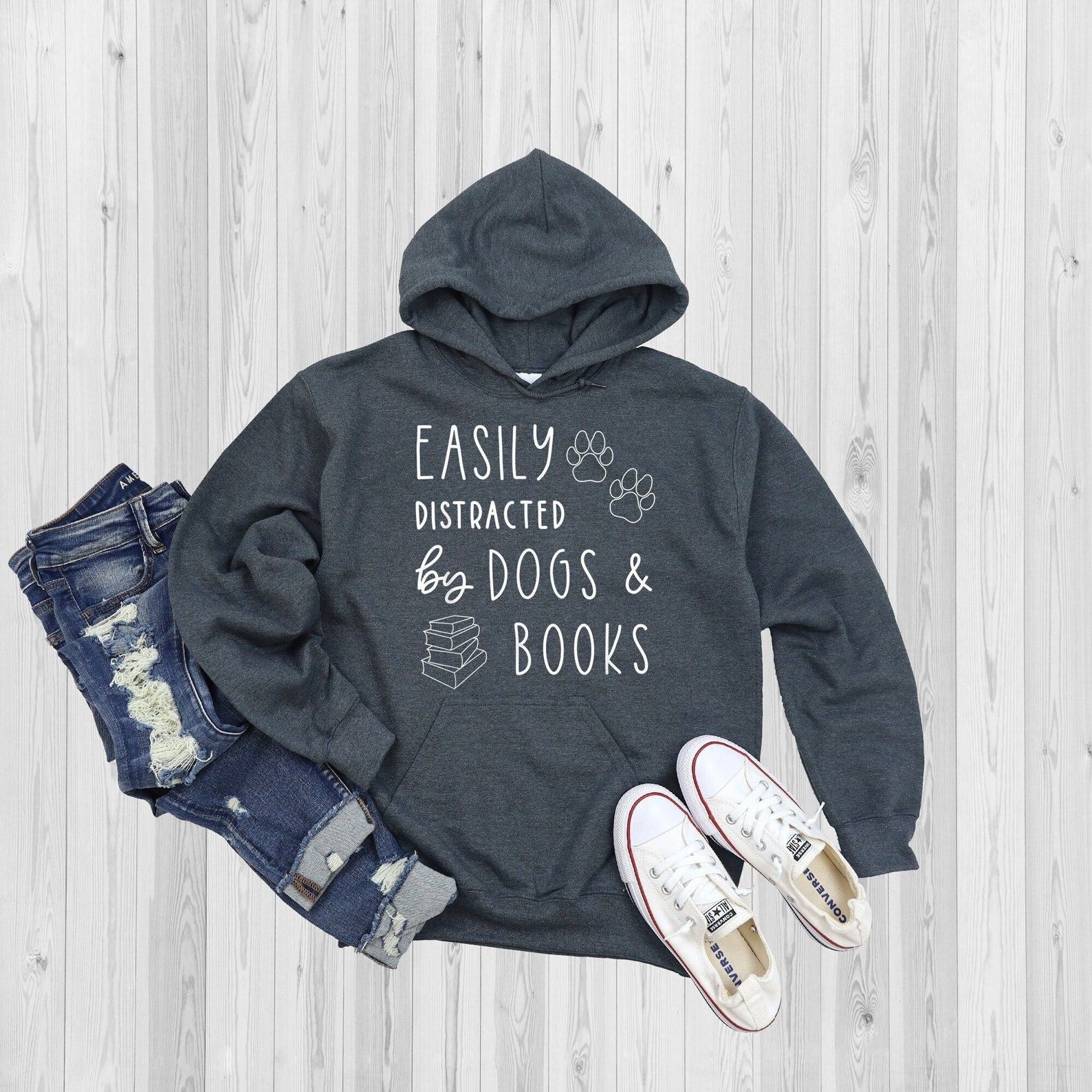 Image of a grey hoodie on a white panel background. There are white converse shoes and jeans beside it. The hoodie has white font that reads "easily distracted by dogs and books." 