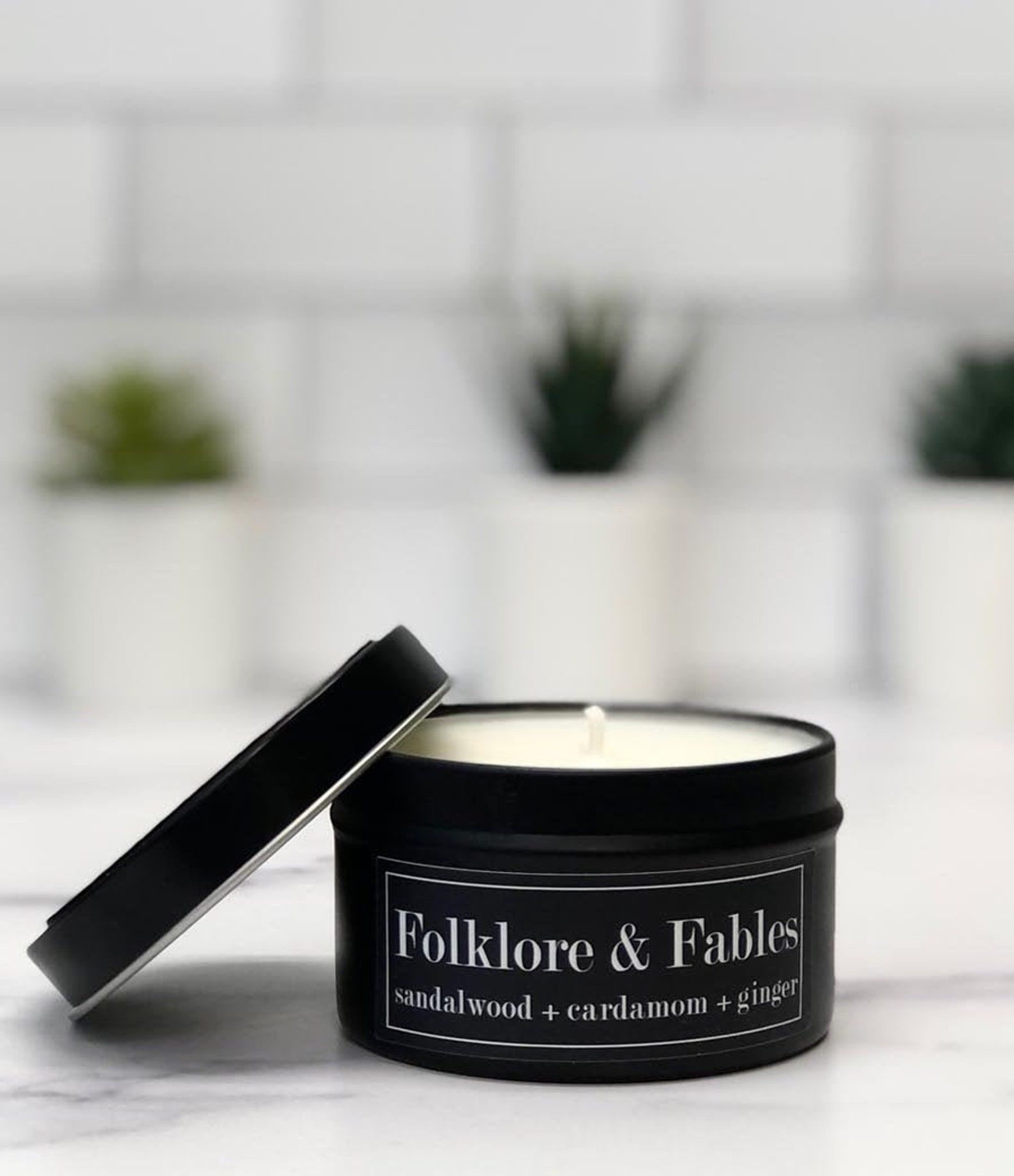 Image of a black candle tin. It reads in white font "Folklore + Fables: Sandalwood + cardamom + ginger." 