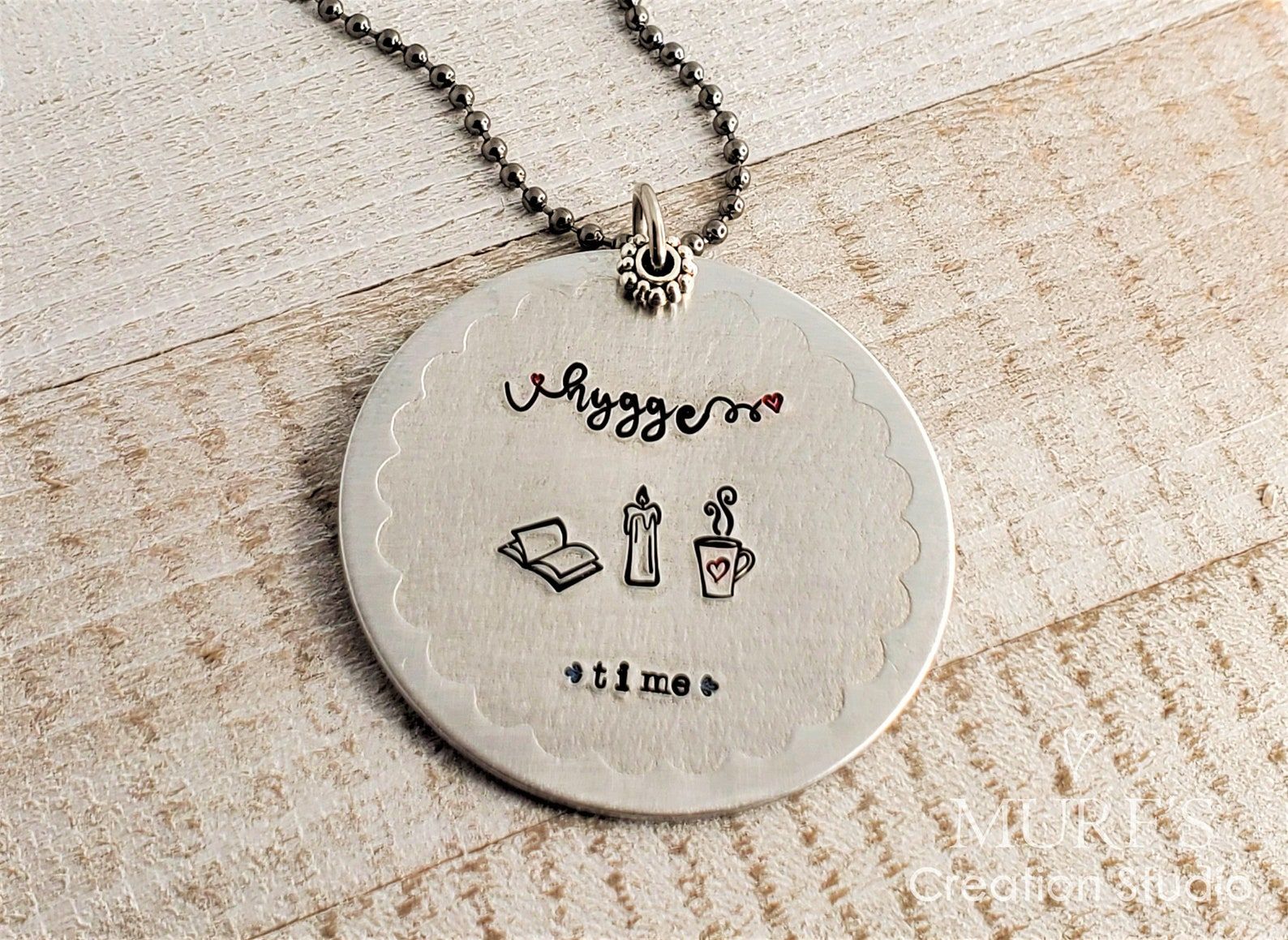 Image of a silver necklace pendant. It features the words "hygge time" and a book, candle, and mug with a warm beverage inside. 