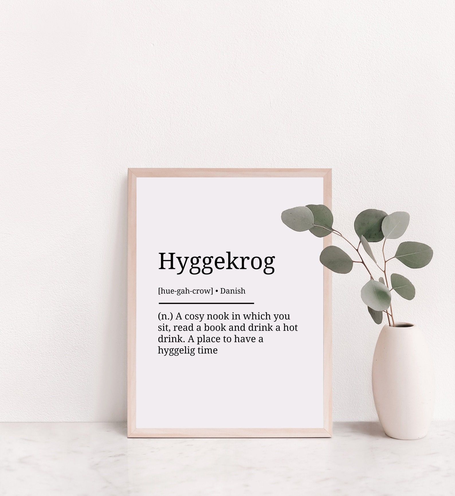 Image of a white print in a white frame. The print reads: "Hyggekrog: A cozy reading nook in which you sit, read a book, and drink a hot drink. A place to have hyggelig time."