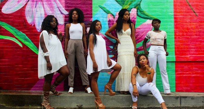 six women of color dressed in light colors standing in front of a colorful mural