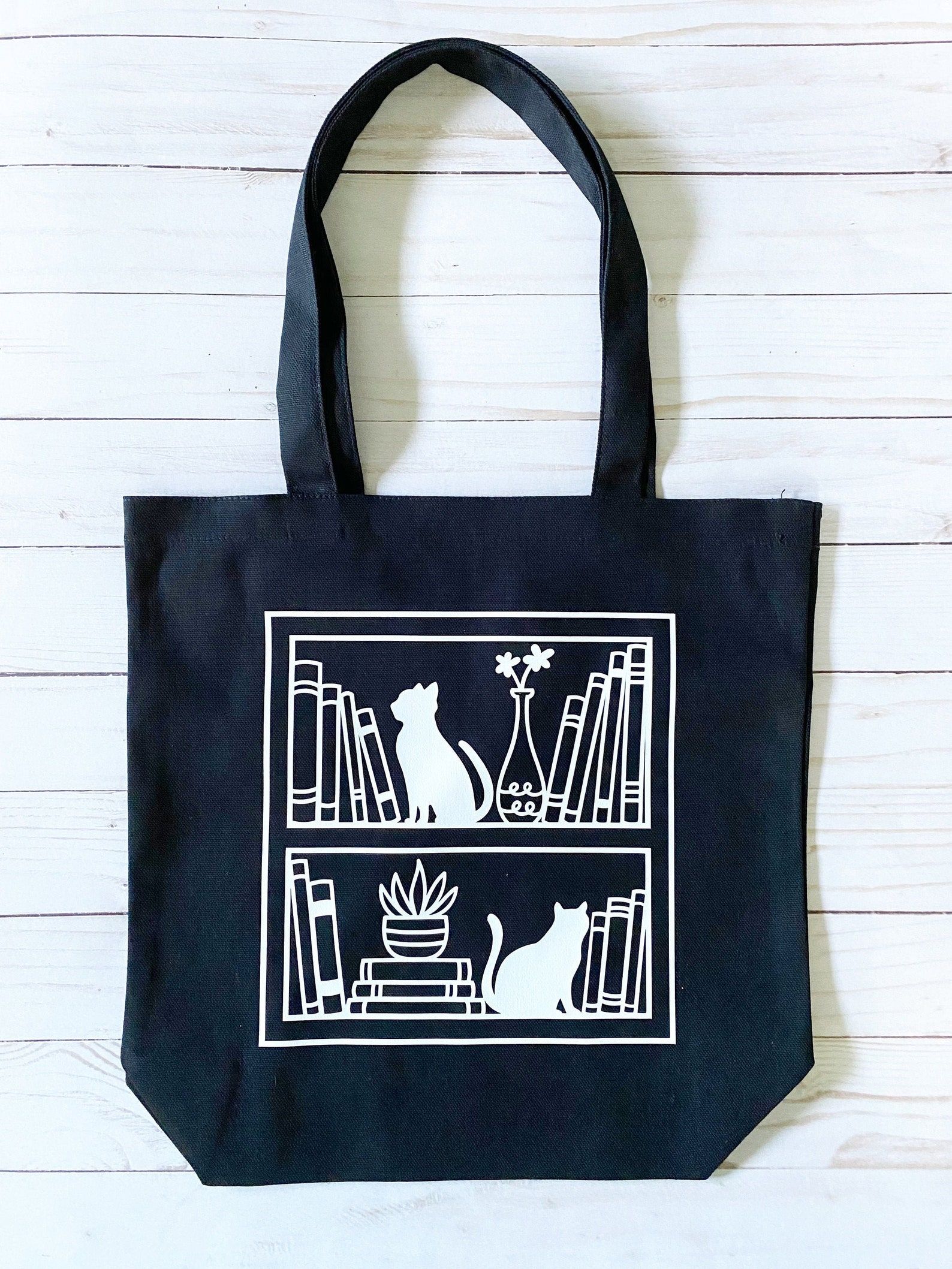 Image d'un sac fourre-tout noir.  L'image blanche représente un ensemble d'étagères avec des livres, ainsi que des chats. 