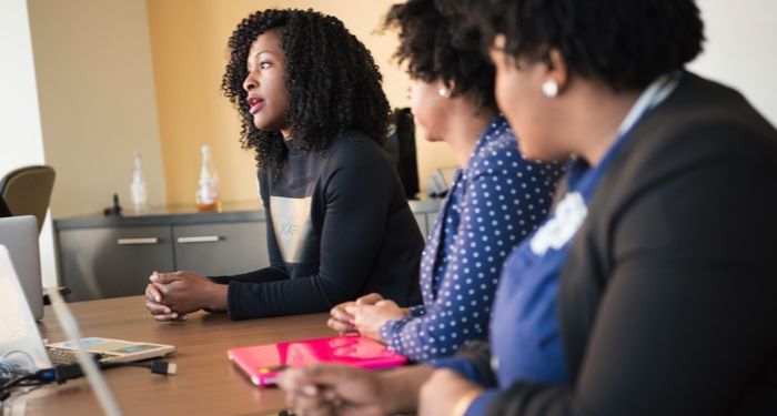 three women seated at a conference table in a meeting with laptop computers and books