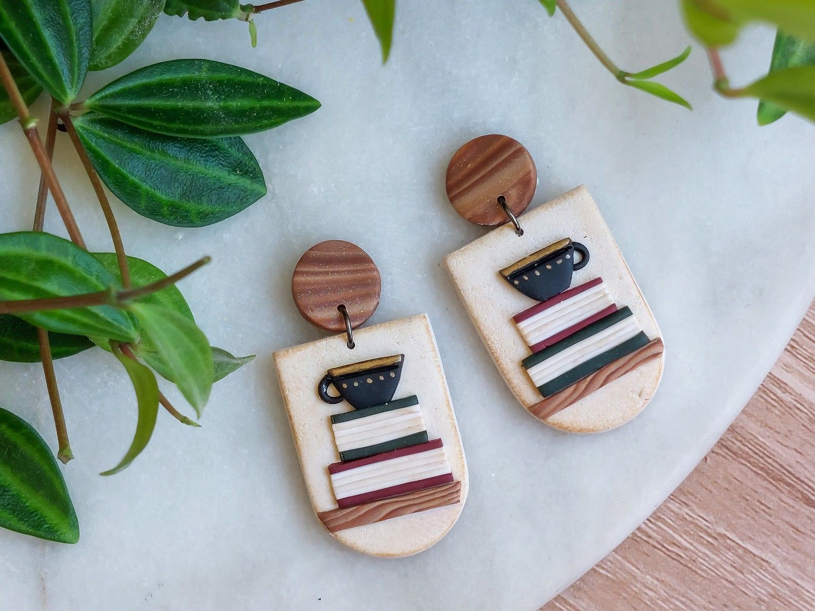 Image of a pair of polymer clay earrings. They have a cream colored base, with two books on them in red and green. On top of the books is a dark blue mug. 