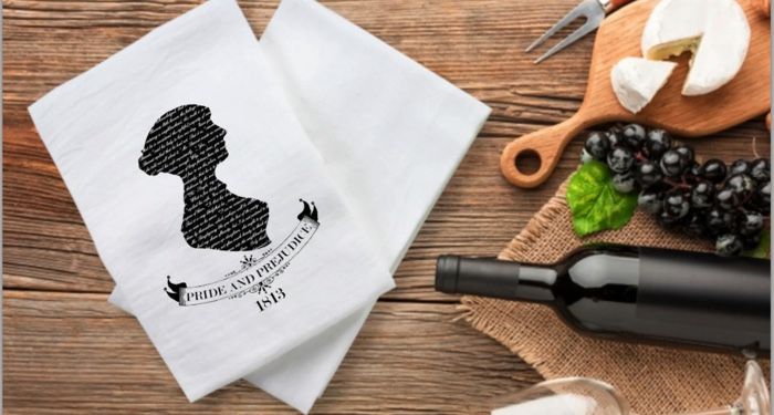 An overhead photo of two white tea towels with a silhouette of a woman in black and the title, "Pride and Prejudice" with the date 1813 below it. To the right of the tea towels is a bottle of wine, glasses, grapes, and a cheese board.