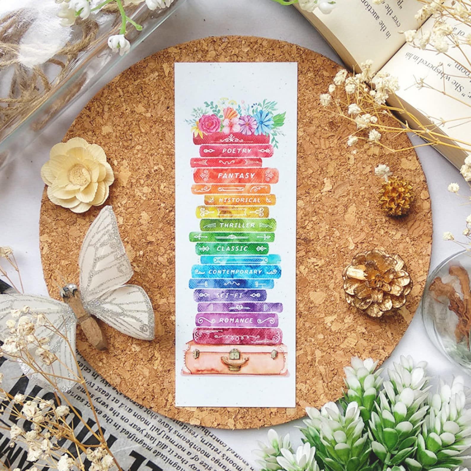 a bookmark depicting a floral book stack in rainbow hues, and with different genres on each book's spine