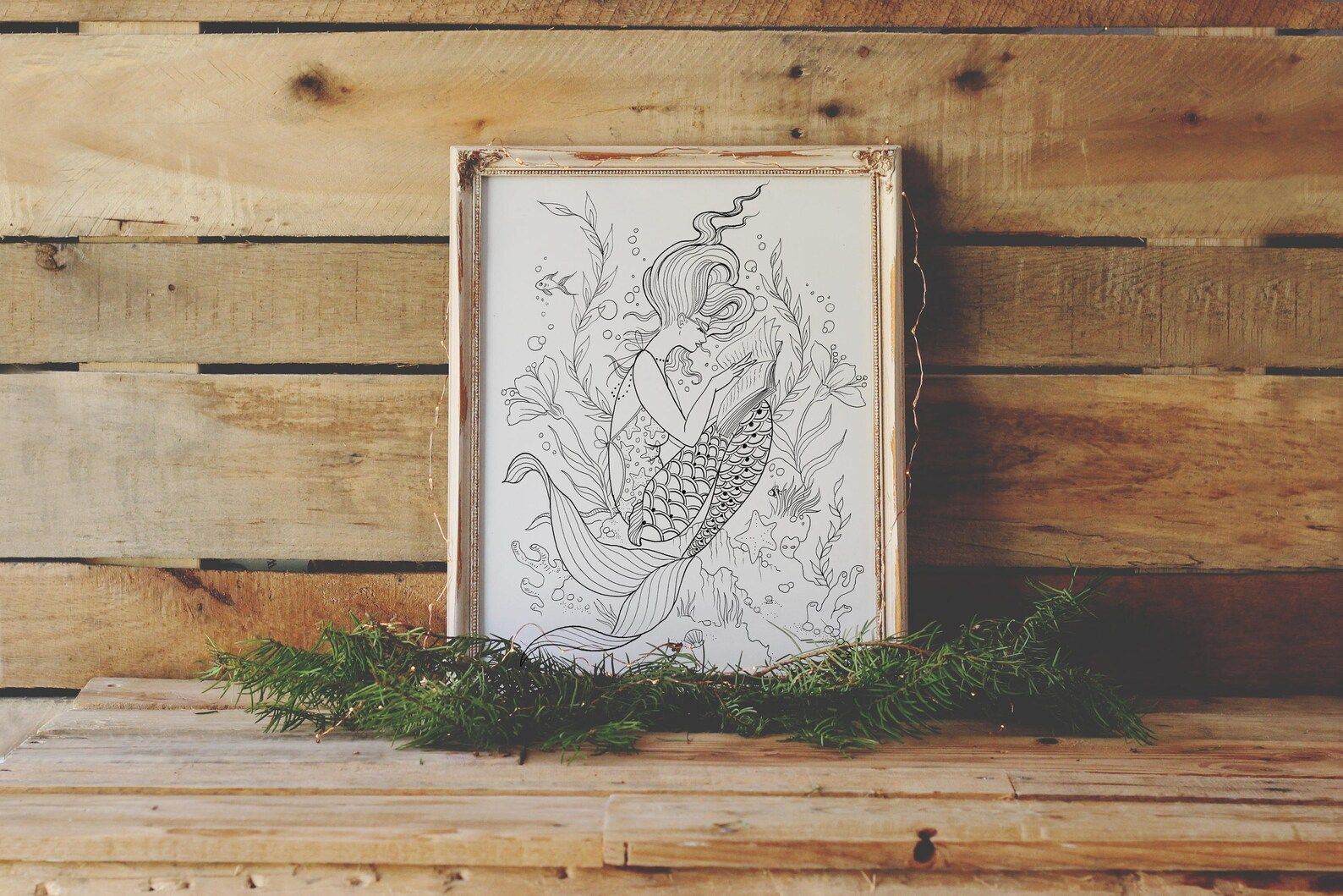 A framed black and white print of a mermaid reading, which can be colored in.