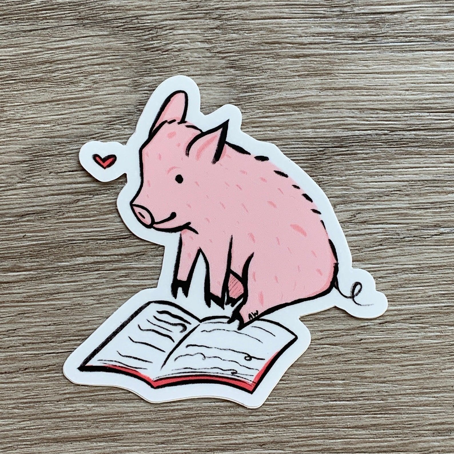 A sticker depicting a hand drawn pink pig reading an open book