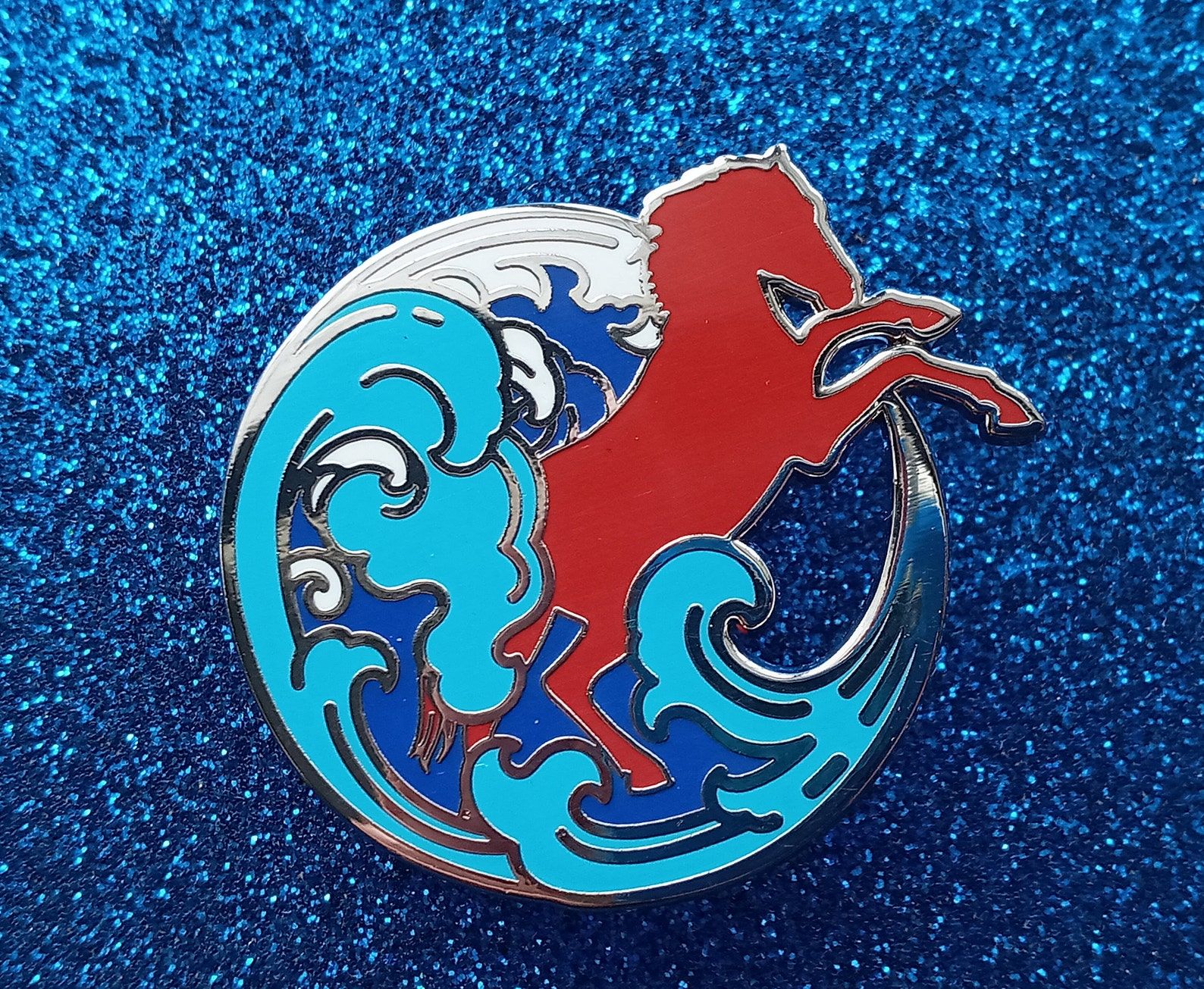 An enamel pin depicting a rearing red horse and blue ocean waves