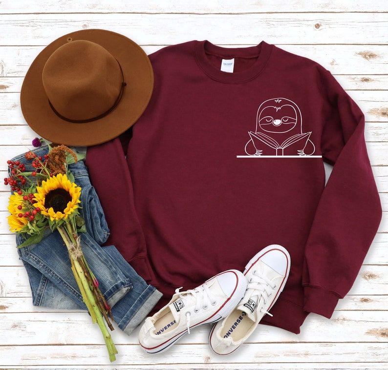 Image of a maroon sweatshirt on a white background. Beside it are a brown hat, white converse shoes, jeans, and a sunflower. The sweatshirt features white ink. There is a sloth with an open book on the left-hand side. 