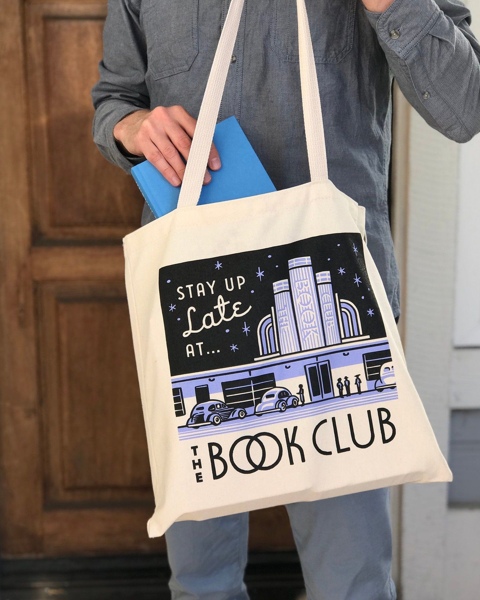 Image of a white person with a tote bag. The canvas bag features an image of a vintage movie theater and the words "Stay up late at the book club." 