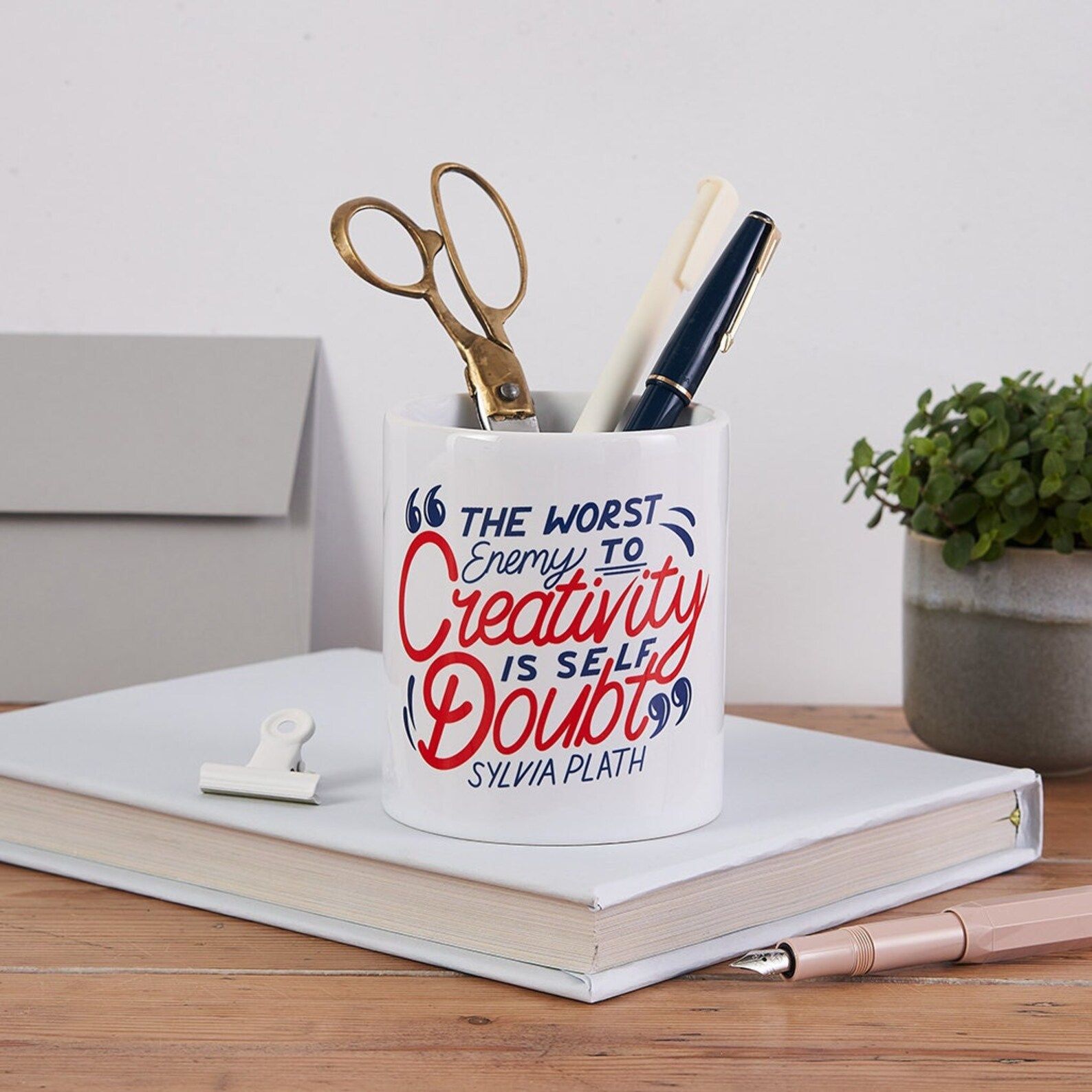 A ceramic pen pot with a quote that reads "The enemy to creativity is Self Doubt. --Sylvia Plath" in red and blue script.