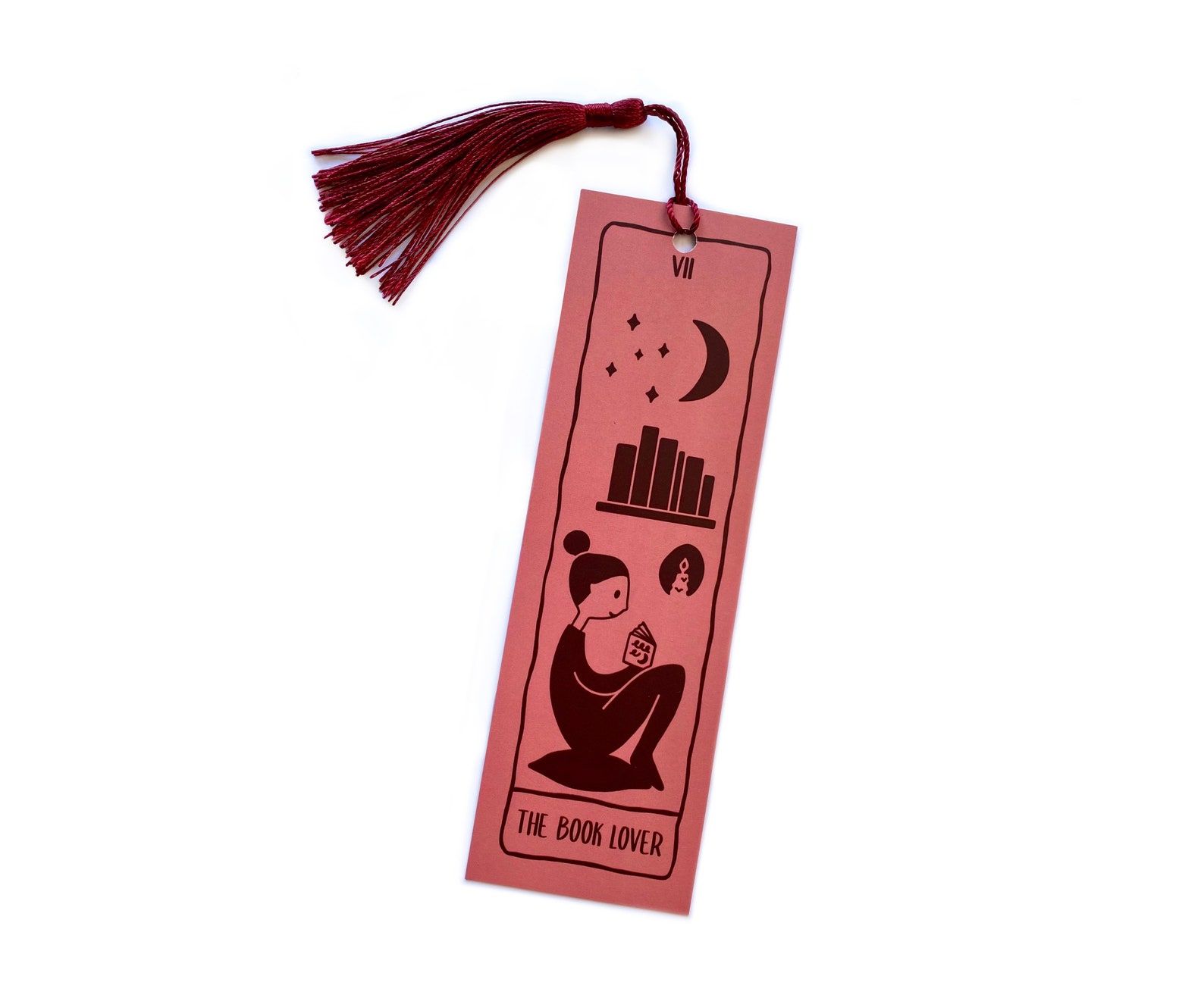 Image of a pink bookmark with a pink tassel. The bookmark is designed like a tarot card called "The Book Lover," featuring a person reading, a bookshelf, and a moon and stars.