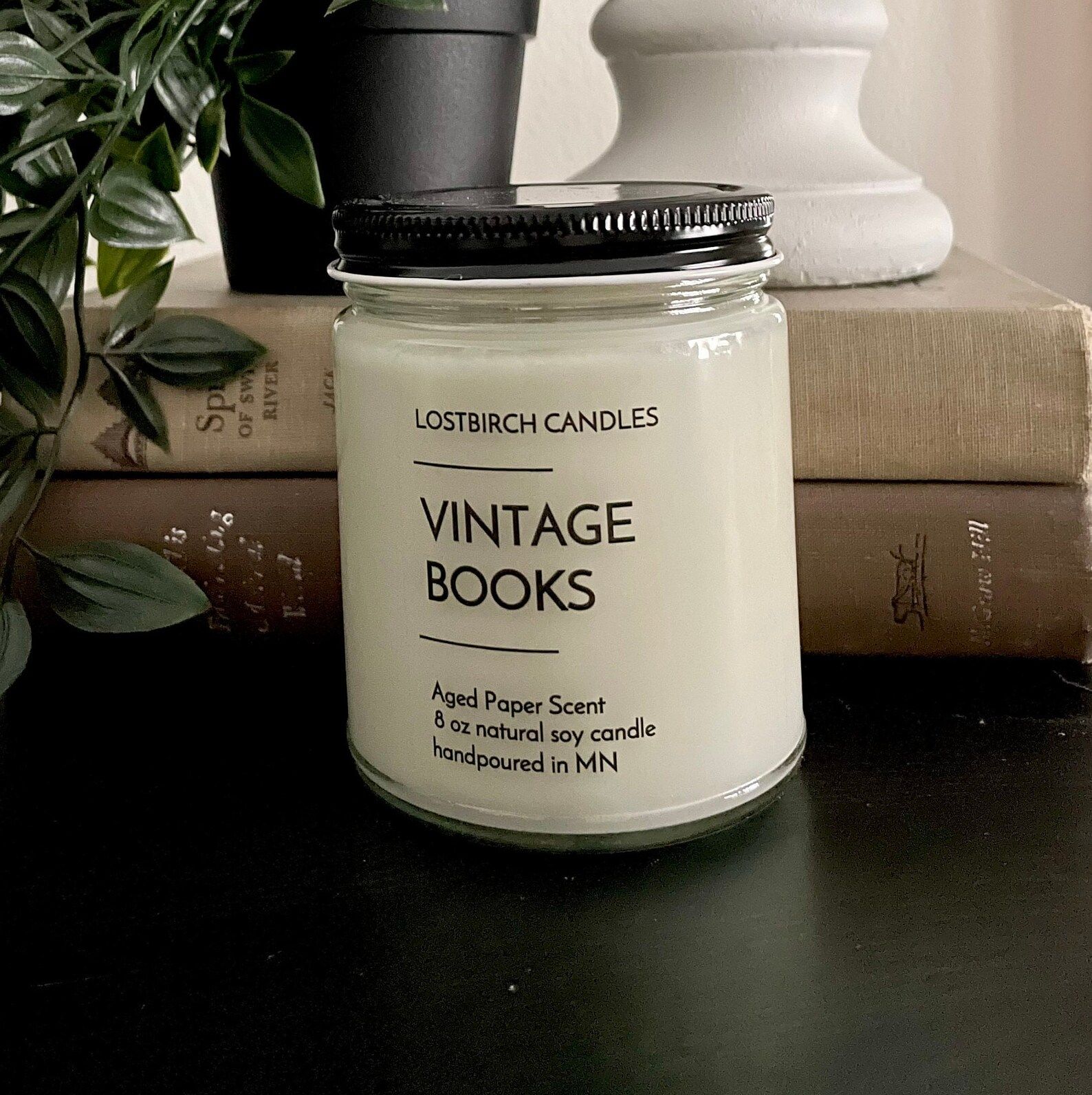 White candle in a glass jar. The har reads "Vintage Books: Aged paper scent." It's on a black table in front of old books. 