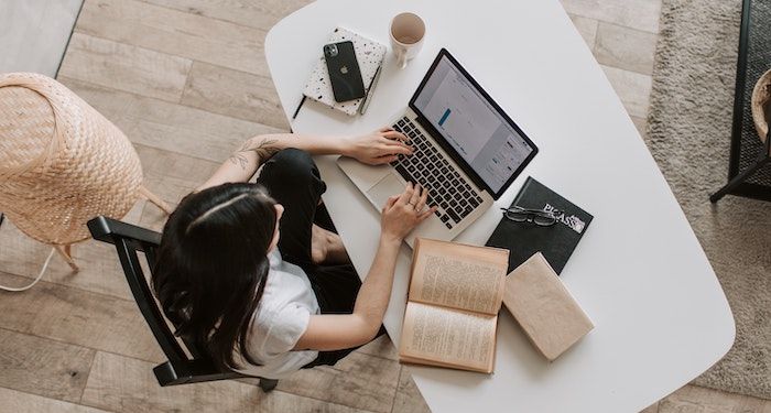 woman writing at laptop with book open beside her