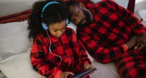 Black man and his daughter laying on the bed while she looks at a tablet and he sleeps