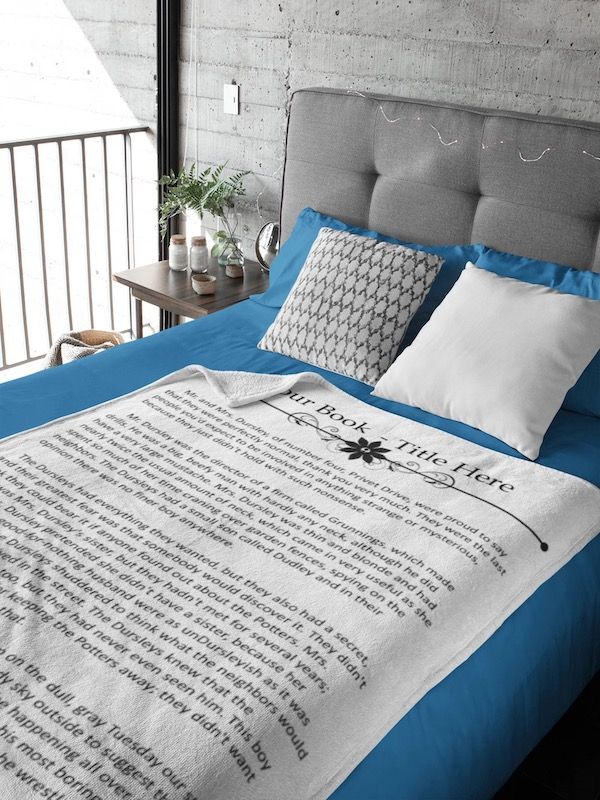 a white blanket with black lettering of text from a book an the words "Your Book Title Here" at the top. The blanket is spread out on top of a bed with a blue comforter