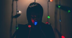 a dark image of a person in a hoodie sweatshirt standing behind multi colored Christmas lights