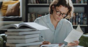 a person seated in front of a bookshelf holding a book in one hand and taking notes with the other, off to the side is a large stack of books