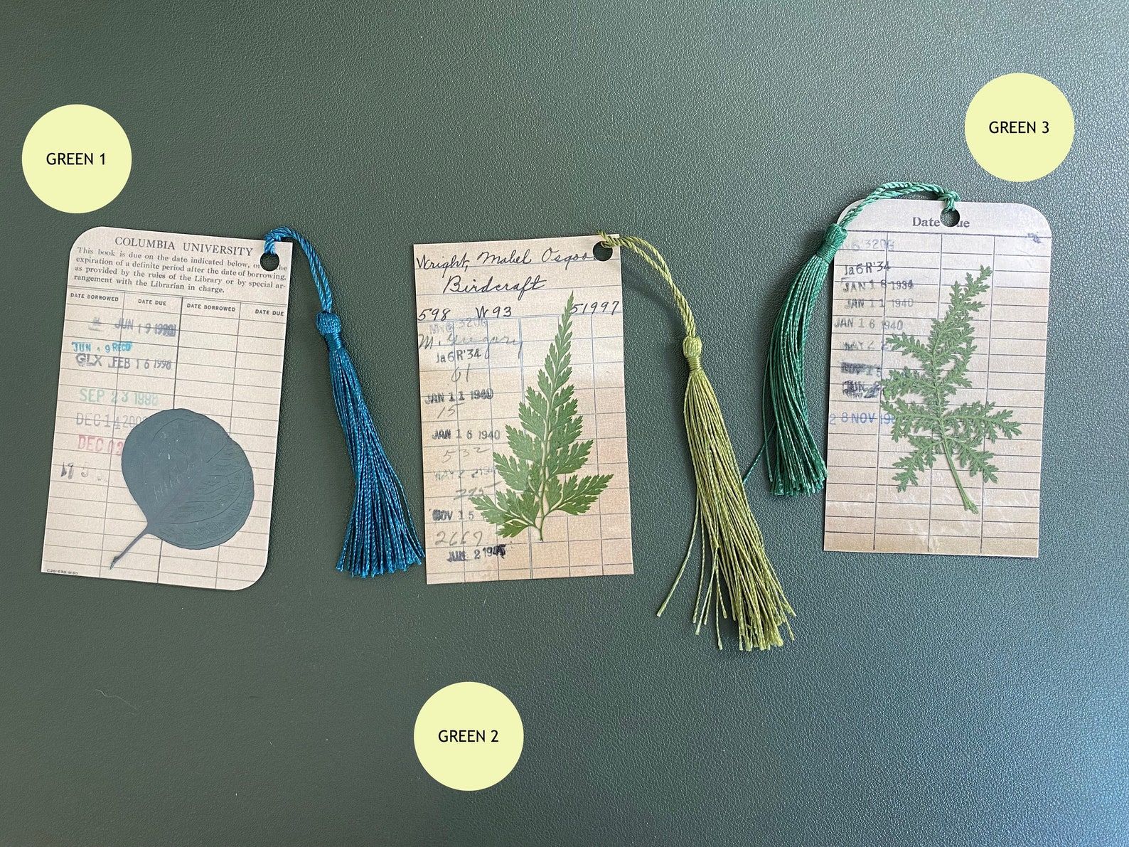 Image of three bookmarks made from vintage library due date cards. Each of the bookmarks has a tassel and a pressed leaf in the center. 