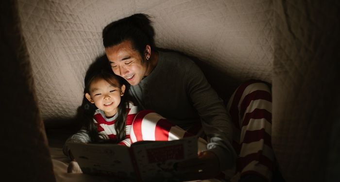 Asian man and his daughter reading a book with a flashlight under a blanket fort and smiling