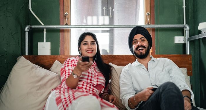 an Indian woman and man sitting on the couch watching tv and smiling