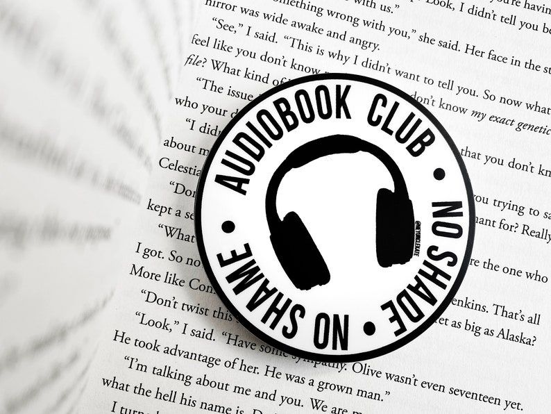 Image of a sticker on an open book. The round sticker has headphones in the center and reads "audiobook club: no shade, no shame" around them. 