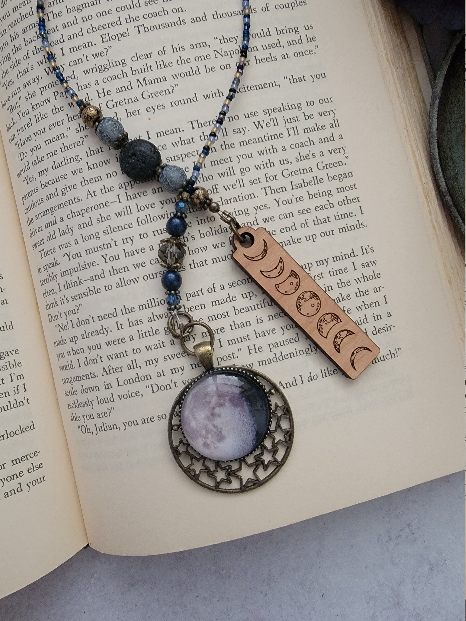 Image of a beaded bookmark. It sits on an open book. One side of the bookmark has a full moon, and the other side of the bookmark has the moon phases. 