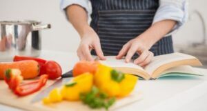 person in an apron thumbing through a cookbook. On the counter in front of them is a cutting board topped with red and yellow bell peppers