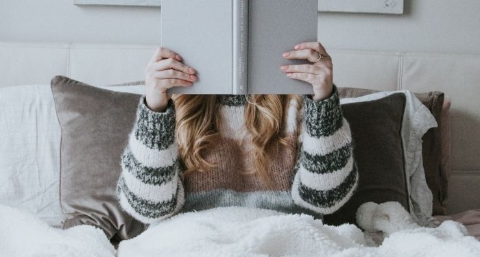 pale skinned person with long blonde hair sits in bed with blankets pulled up while holding a book up and obscuring their face