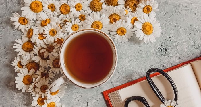 a cup of tea with daisies surrounding one side of it, and an open book with a pair of scissors sitting on top