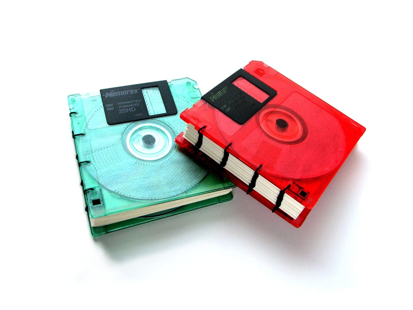 Two small notebooks on a white background. The notebook on the left is made from a green floppy disk and the one on the right is red. 