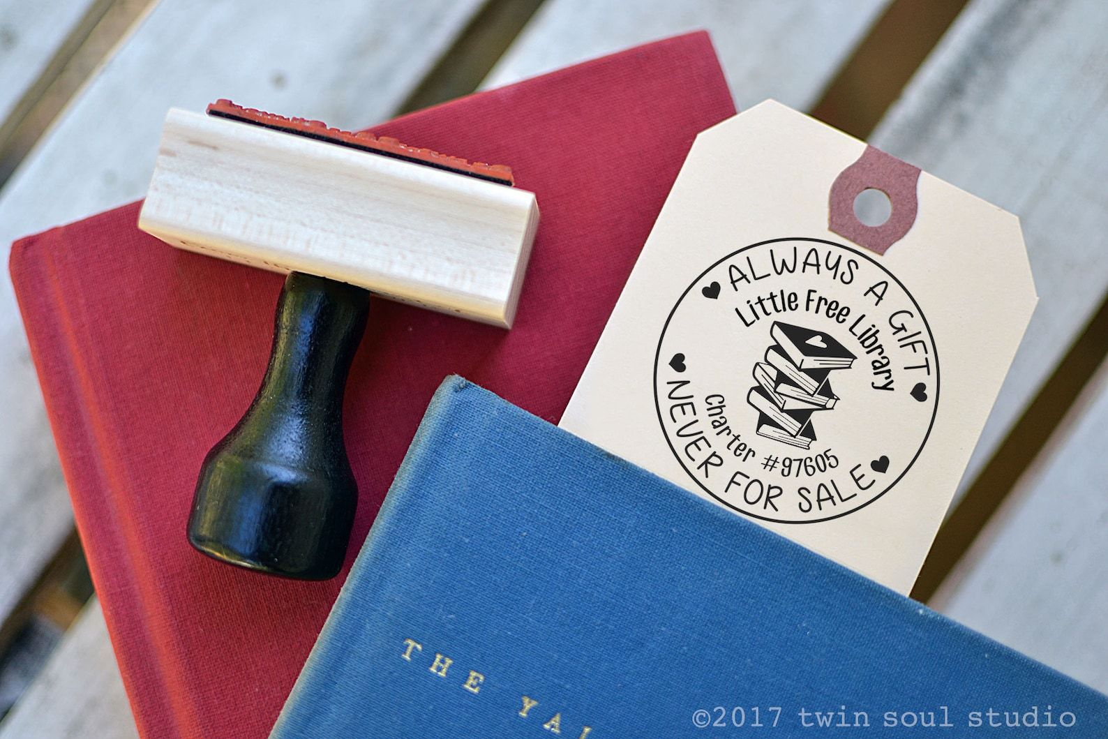 A stamp on a cream tag that reads "Always a Gift, Never for Sale: Little Free Library Charter # XXXXXX" with a stack of books